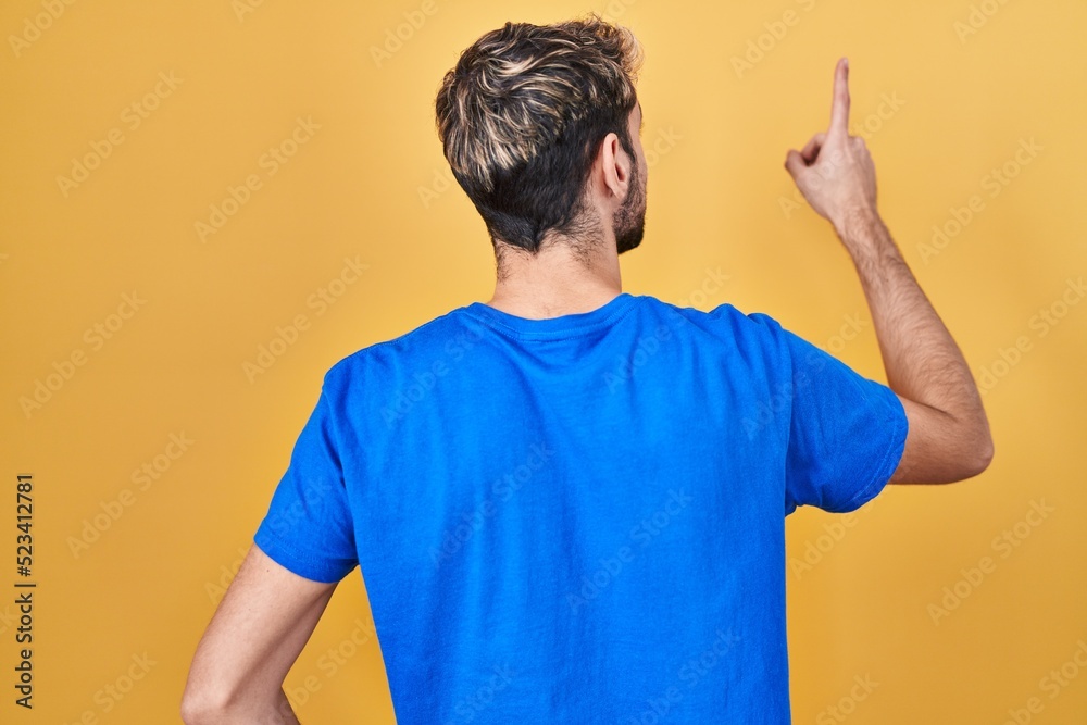 Hispanic man with beard standing over yellow background posing backwards pointing ahead with finger hand Hispanic man with beard standing over yellow background posing backwards pointing ahead with finger hand