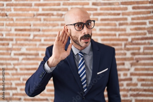 Bald man with beard wearing business clothes and glasses smiling with hand over ear listening an hearing to rumor or gossip. deafness concept.