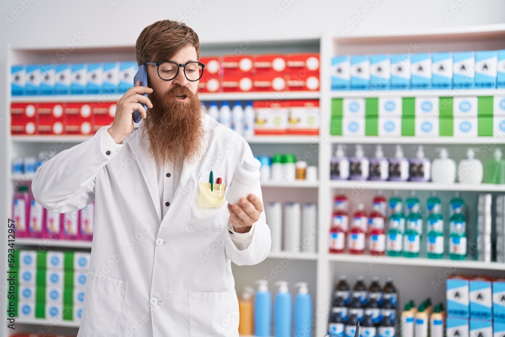 Young redhead man pharmacist holding pills bottle talking on smartphone at pharmacy Young redhead man pharmacist holding pills bottle talking on smartphone at pharmacy