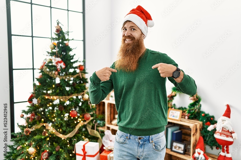 Redhead man with long beard wearing christmas hat by christmas tree looking confident with smile on face, pointing oneself with fingers proud and happy.