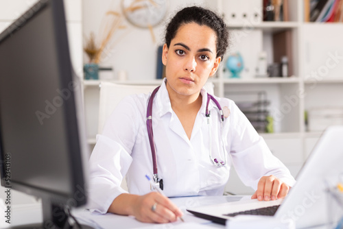 Young woman doctor assistant working in medical office using laptop computer and writing prescription