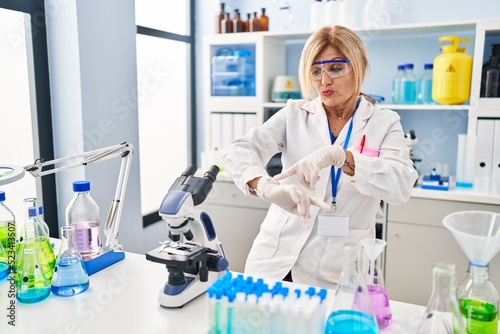 Middle age blonde woman working at scientist laboratory in hurry pointing to watch time  impatience  upset and angry for deadline delay