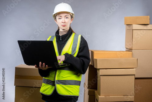 Successful warehouse worker. Girl in logistics warehouse. Fulfillment manager woman. Warehouse worker stands with laptop. Lots of boxes behind businesswoman. Logistic girl in yellow vest