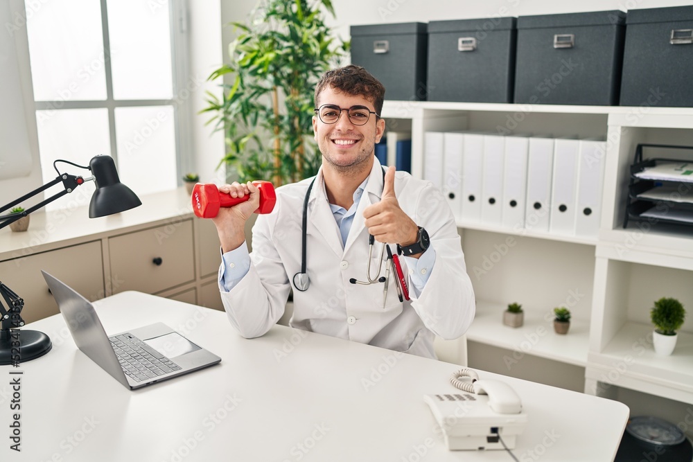 Young hispanic doctor man holding dumbbells smiling happy and positive, thumb up doing excellent and approval sign
