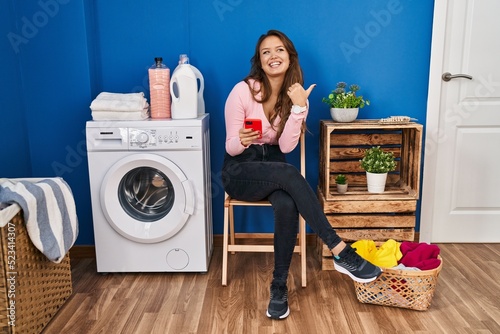 Young hispanic woman sitting waiting for laundry using smartphone smiling with happy face looking and pointing to the side with thumb up.