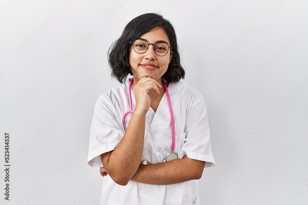 Young hispanic doctor woman wearing stethoscope over isolated background looking confident at the camera smiling with crossed arms and hand raised on chin. thinking positive. Young hispanic doctor woman wearing stethoscope over isolated background looking confident at the camera smiling with crossed arms and hand raised on chin. thinking positive.