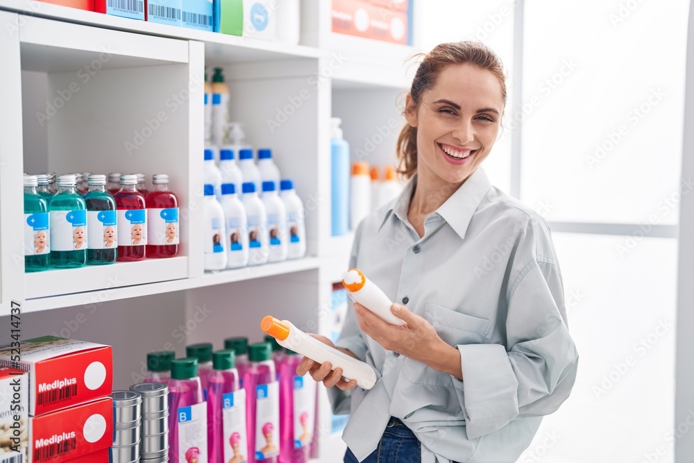 Young woman customer smiling confident holding sunscreen bottles at pharmacy