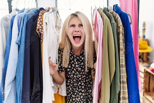 Young blonde woman searching clothes on clothing rack angry and mad screaming frustrated and furious, shouting with anger looking up.