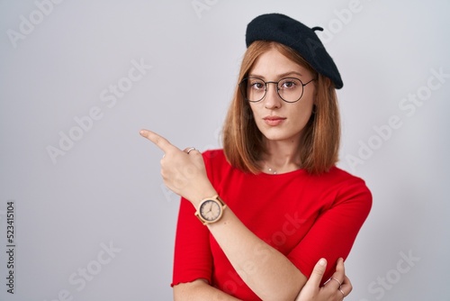 Young redhead woman standing wearing glasses and beret pointing with hand finger to the side showing advertisement, serious and calm face