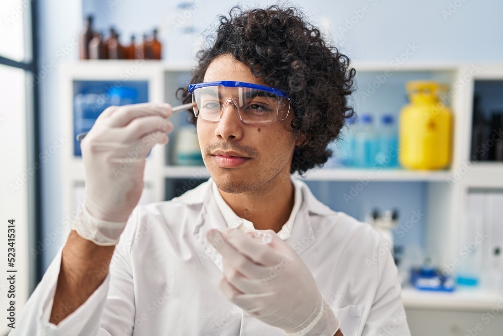 Young hispanic man wearing scientist uniform holding sample with tweezer at laboratory Young hispanic man wearing scientist uniform holding sample with tweezer at laboratory