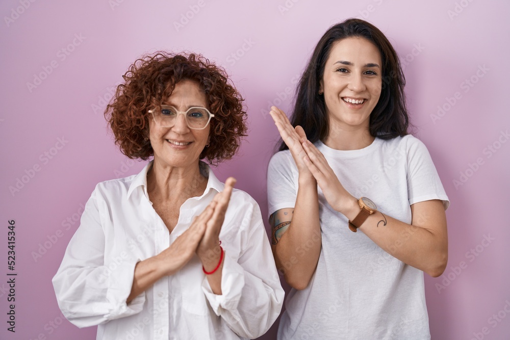 Hispanic mother and daughter wearing casual white t shirt over pink background clapping and applauding happy and joyful, smiling proud hands together Hispanic mother and daughter wearing casual white t shirt over pink background clapping and applauding happy and joyful, smiling proud hands together