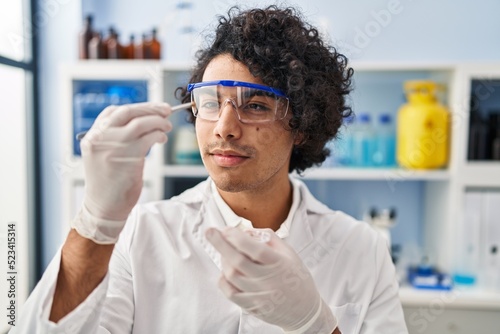 Young hispanic man wearing scientist uniform holding sample with tweezer at laboratory