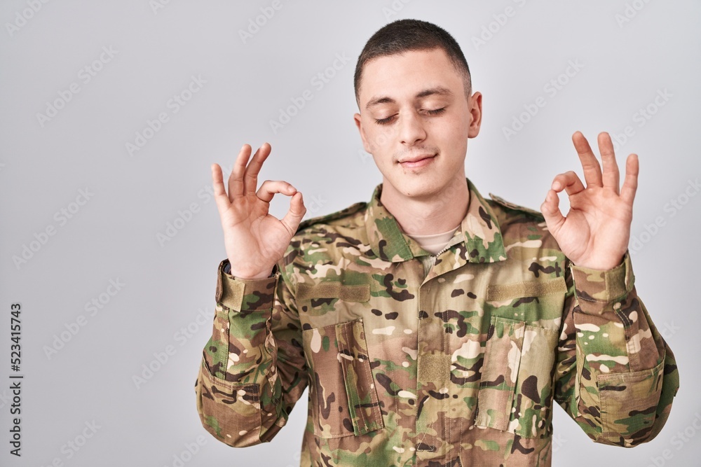 Young man wearing camouflage army uniform relax and smiling with eyes closed doing meditation gesture with fingers. yoga concept. Young man wearing camouflage army uniform relax and smiling with eyes closed doing meditation gesture with fingers. yoga concept.