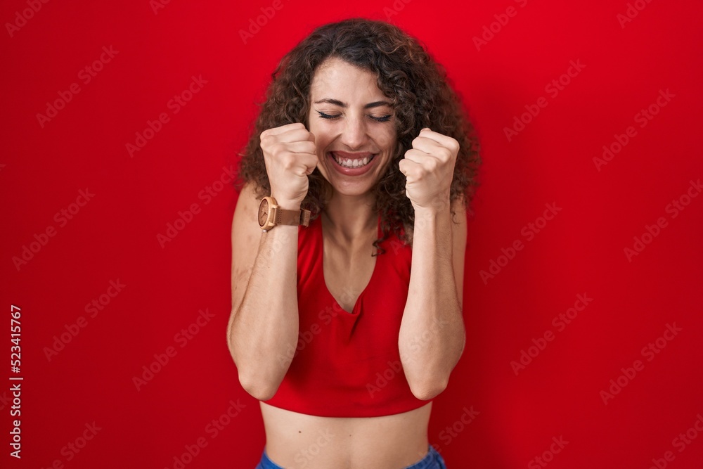Hispanic woman with curly hair standing over red background excited for success with arms raised and eyes closed celebrating victory smiling. winner concept. Hispanic woman with curly hair standing over red background excited for success with arms raised and eyes closed celebrating victory smiling. winner concept.