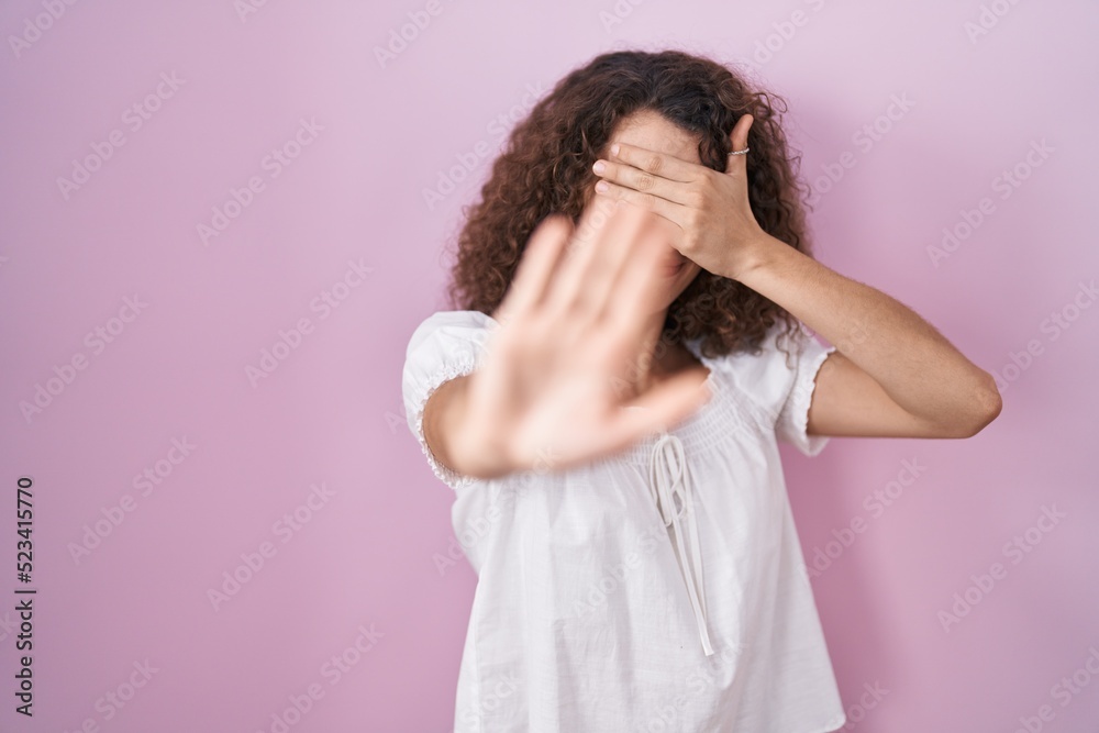 Hispanic woman with curly hair standing over pink background covering eyes with hands and doing stop gesture with sad and fear expression. embarrassed and negative concept. Hispanic woman with curly hair standing over pink background covering eyes with hands and doing stop gesture with sad and fear expression. embarrassed and negative concept.