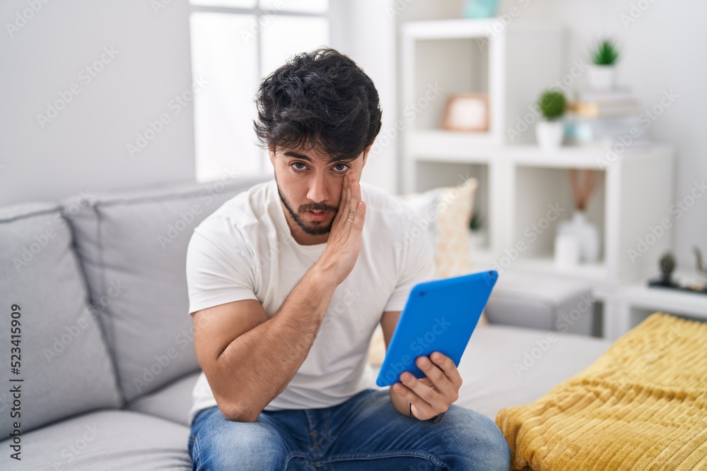 Hispanic man with beard using touchpad sitting on the sofa hand on mouth telling secret rumor, whispering malicious talk conversation Hispanic man with beard using touchpad sitting on the sofa hand on mouth telling secret rumor, whispering malicious talk conversation