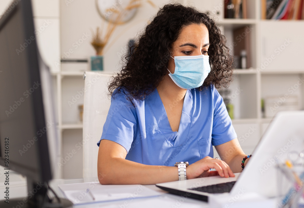 Attentively young female doctor in protective mask working at laptop in her office