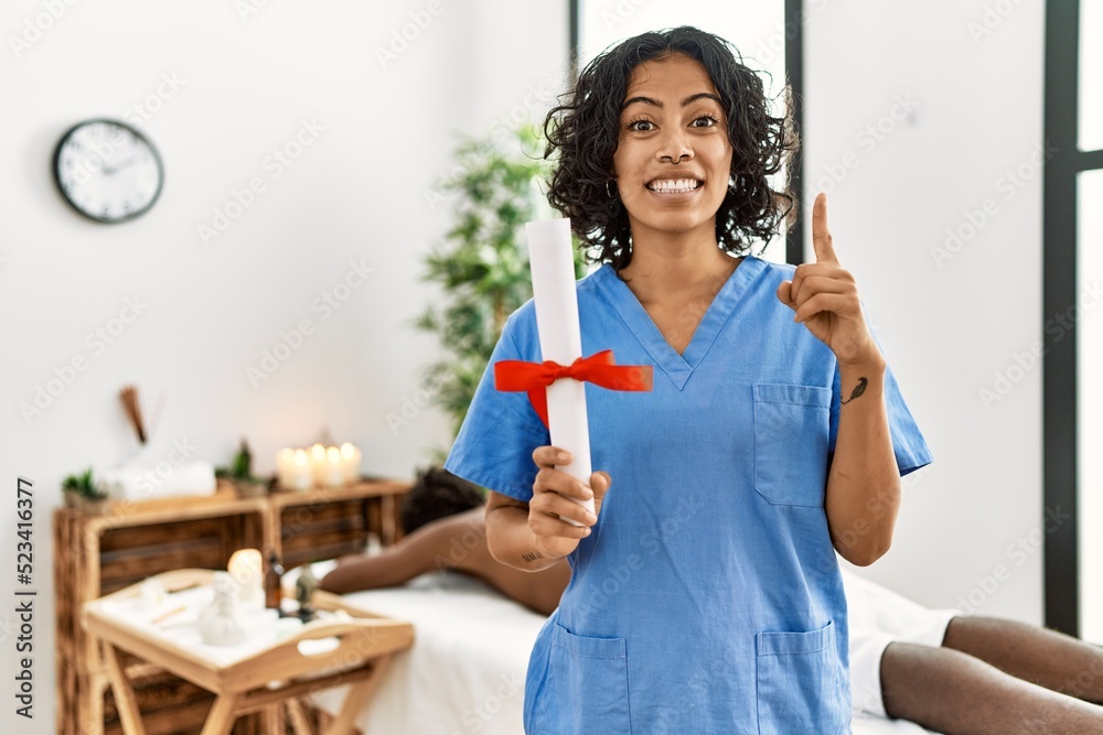 Young therapist woman holding diploma at spa wellness center surprised with an idea or question pointing finger with happy face, number one Young therapist woman holding diploma at spa wellness center surprised with an idea or question pointing finger with happy face, number one