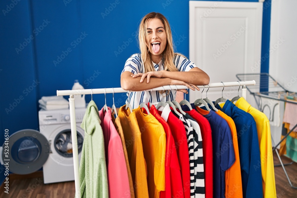 Young blonde woman at laundry room with clean clothes sticking tongue out happy with funny expression.