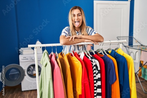 Young blonde woman at laundry room with clean clothes sticking tongue out happy with funny expression.