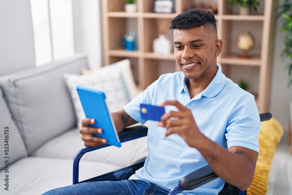 Young latin man using touchpad and credit card sitting on wheelchair at home Young latin man using touchpad and credit card sitting on wheelchair at home