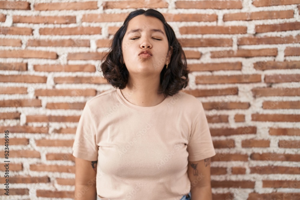 Young hispanic woman standing over bricks wall looking at the camera blowing a kiss on air being lovely and sexy. love expression. Young hispanic woman standing over bricks wall looking at the camera blowing a kiss on air being lovely and sexy. love expression.