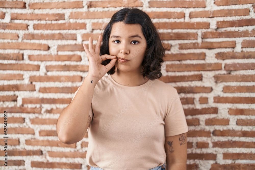 Young hispanic woman standing over bricks wall mouth and lips shut as zip with fingers. secret and silent, taboo talking Young hispanic woman standing over bricks wall mouth and lips shut as zip with fingers. secret and silent, taboo talking