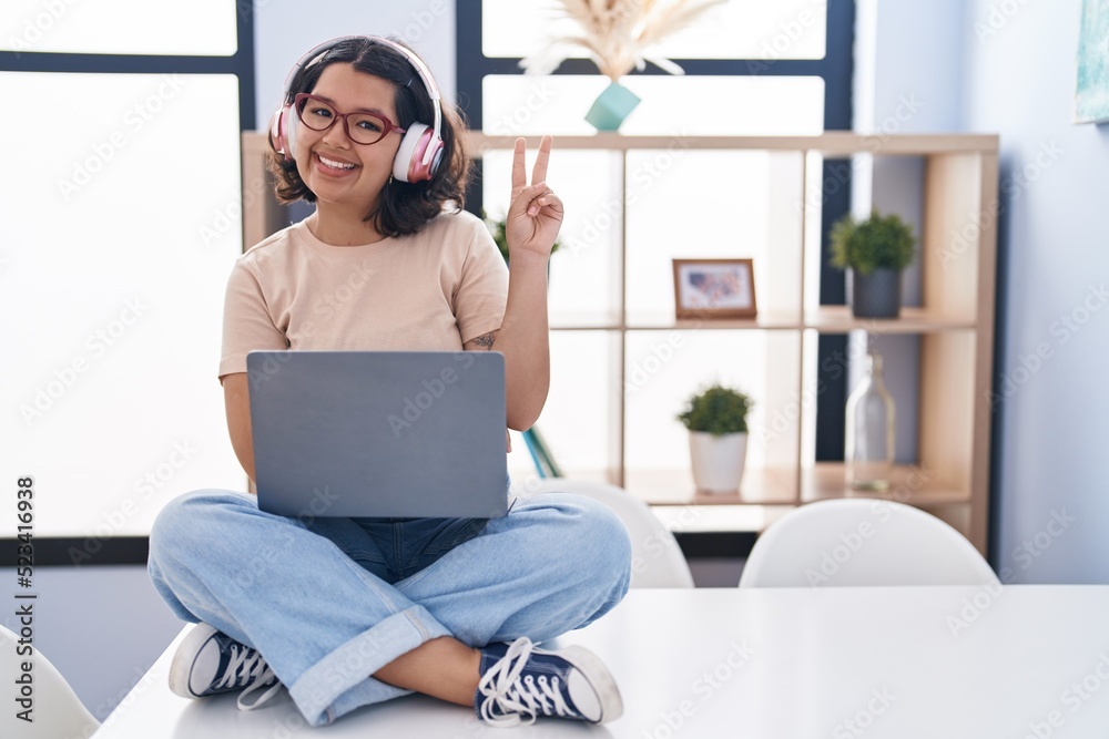 Young hispanic woman using laptop sitting on the table wearing headphones smiling with happy face winking at the camera doing victory sign. number two. Young hispanic woman using laptop sitting on the table wearing headphones smiling with happy face winking at the camera doing victory sign. number two.