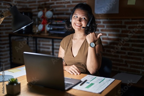 Young hispanic woman working at the office at night smiling with happy face looking and pointing to the side with thumb up.
