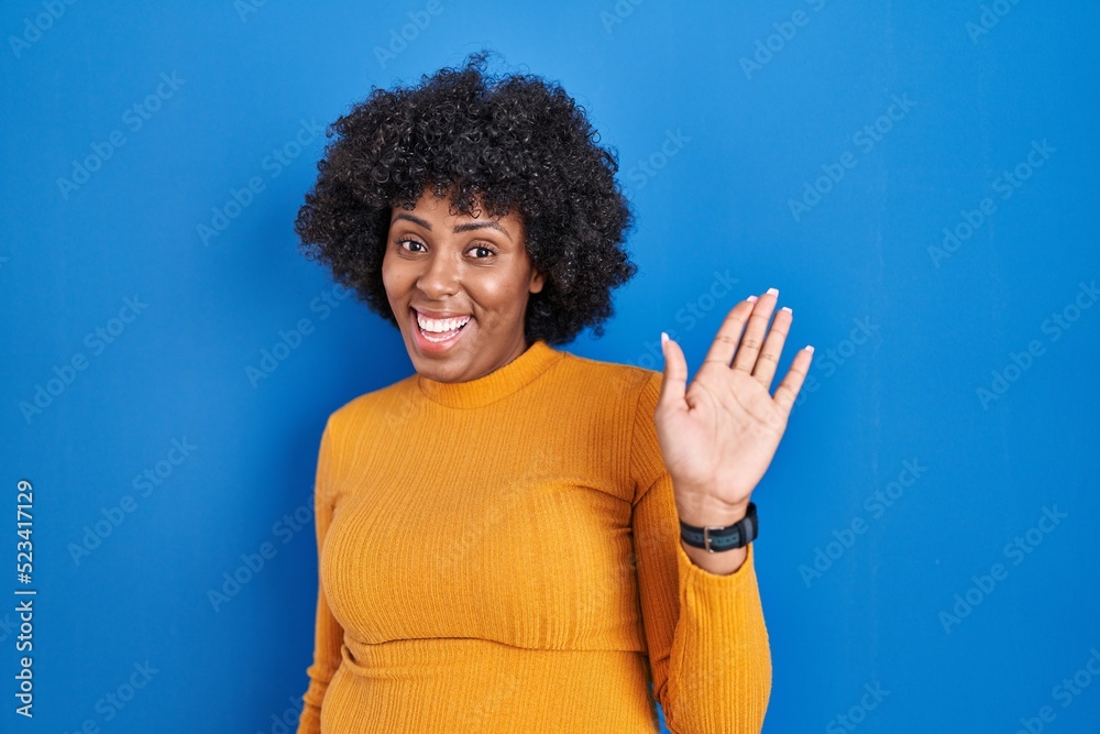 Black woman with curly hair standing over blue background waiving saying hello happy and smiling, friendly welcome gesture Black woman with curly hair standing over blue background waiving saying hello happy and smiling, friendly welcome gesture
