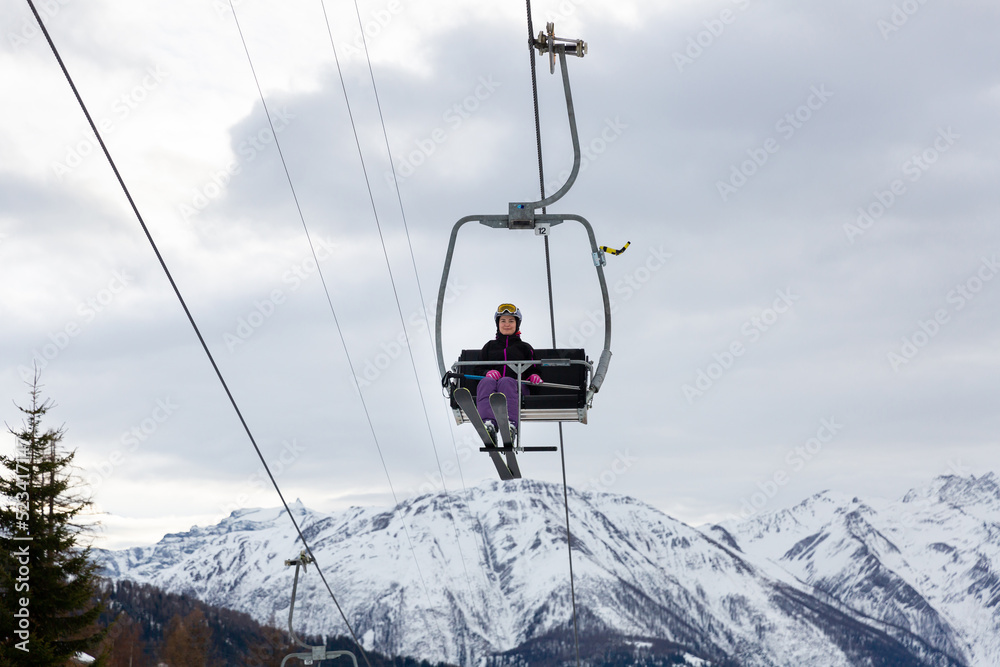 Smiling woman skier in full ski equipment riding on chairlift, lifting to top of mountain to pistes on background of snowy peaks on cloudy winter day..