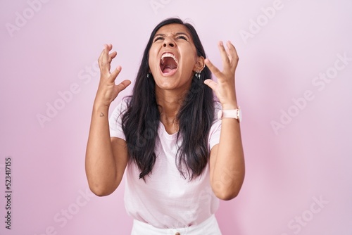 Young hispanic woman standing over pink background crazy and mad shouting and yelling with aggressive expression and arms raised. frustration concept.