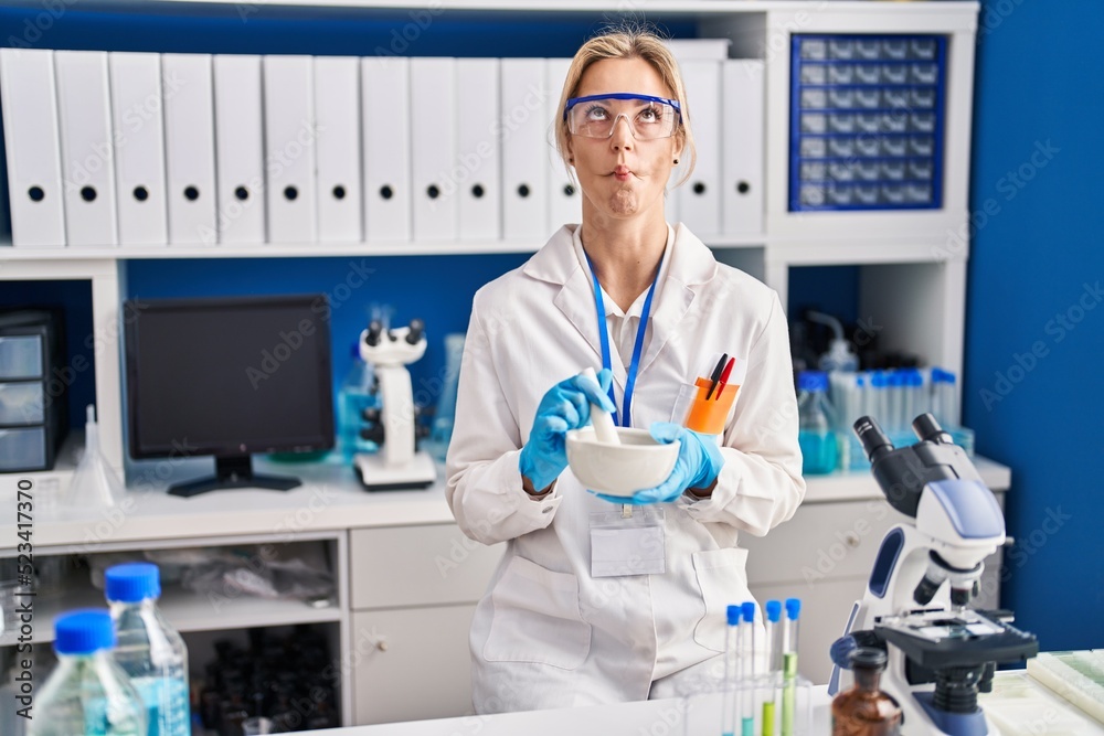 Young caucasian woman working at scientist laboratory making fish face with mouth and squinting eyes, crazy and comical.