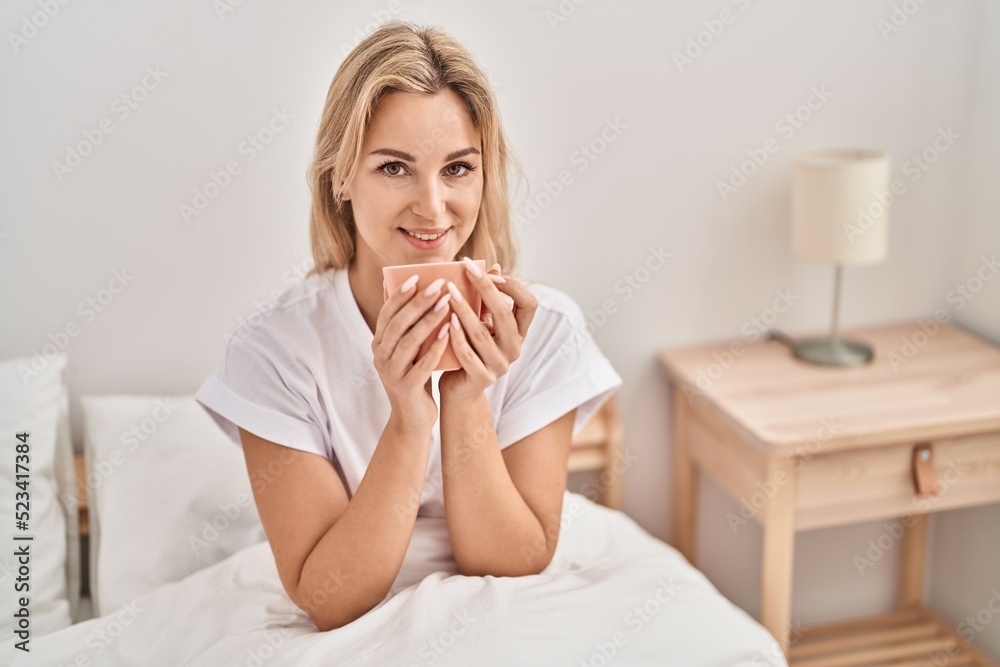 Young blonde woman drinking cup of coffee sitting on bed at bedroom Young blonde woman drinking cup of coffee sitting on bed at bedroom