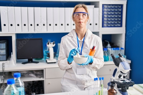 Young caucasian woman working at scientist laboratory making fish face with mouth and squinting eyes  crazy and comical.