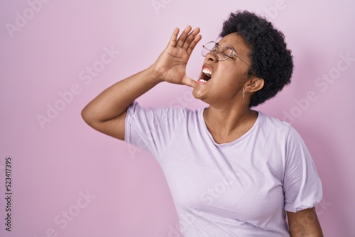 Young african american woman standing over pink background shouting and screaming loud to side with hand on mouth. communication concept.