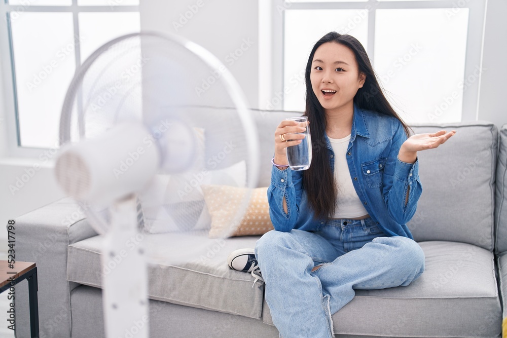 Young asian woman drinking glass of water enjoying air from fan celebrating achievement with happy smile and winner expression with raised hand Young asian woman drinking glass of water enjoying air from fan celebrating achievement with happy smile and winner expression with raised hand