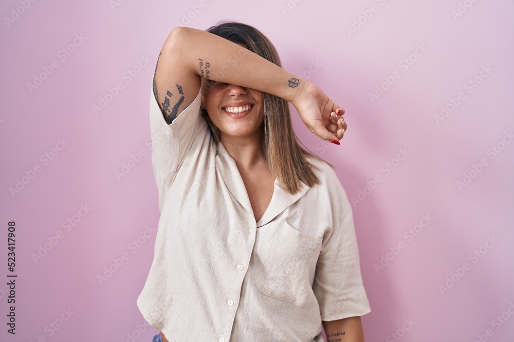 Blonde woman standing over pink background covering eyes with arm smiling cheerful and funny. blind concept.