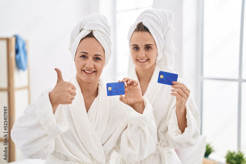 Middle age woman and daughter wearing bathrobe holding credit card smiling happy and positive, thumb up doing excellent and approval sign Middle age woman and daughter wearing bathrobe holding credit card smiling happy and positive, thumb up doing excellent and approval sign