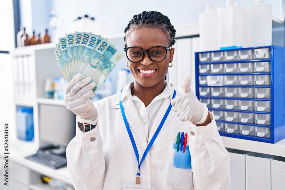African woman with braids working at scientist laboratory holding money smiling happy and positive, thumb up doing excellent and approval sign African woman with braids working at scientist laboratory holding money smiling happy and positive, thumb up doing excellent and approval sign