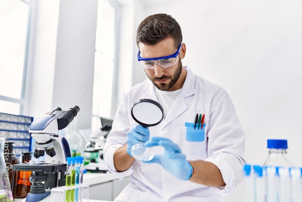 Young hispanic man wearing scientist uniform using loupe at laboratory