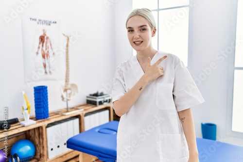 Young caucasian woman working at pain recovery clinic cheerful with a smile on face pointing with hand and finger up to the side with happy and natural expression