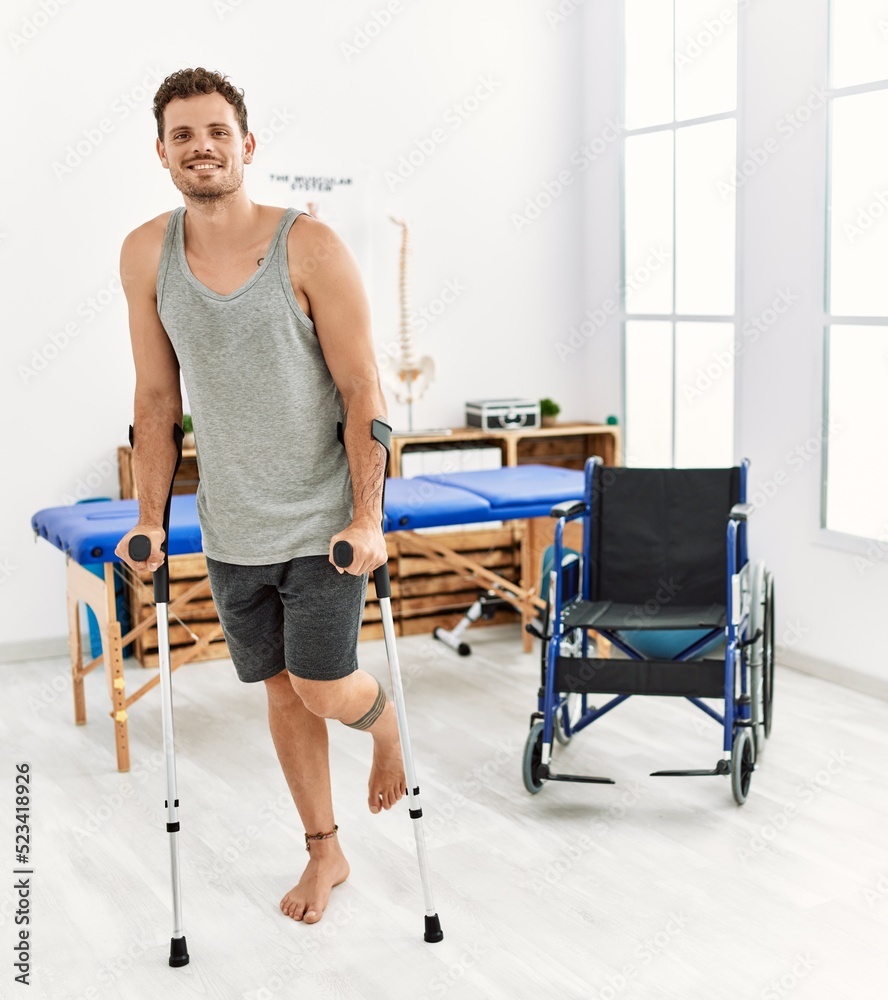 Young hispanic man smiling confident walking using crutches at clinic