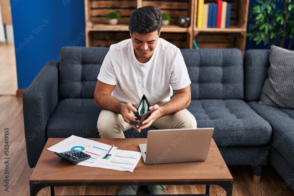 Young hispanic man using laptop holding wallet with dollars at home Young hispanic man using laptop holding wallet with dollars at home