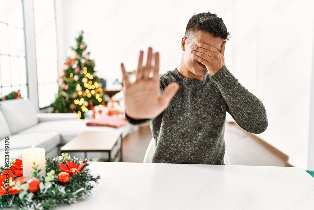 Young hispanic man sitting on the table by christmas tree covering eyes with hands and doing stop gesture with sad and fear expression. embarrassed and negative concept.