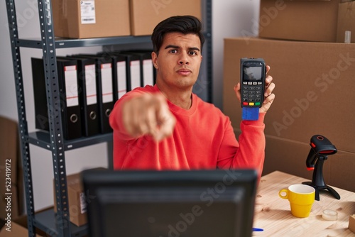 Hispanic man working at small business ecommerce holding dataphone pointing with finger to the camera and to you, confident gesture looking serious photo