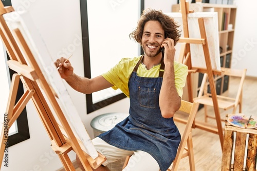 Young hispanic artist man smiling happy drawing and talking on the smartphone at art studio.