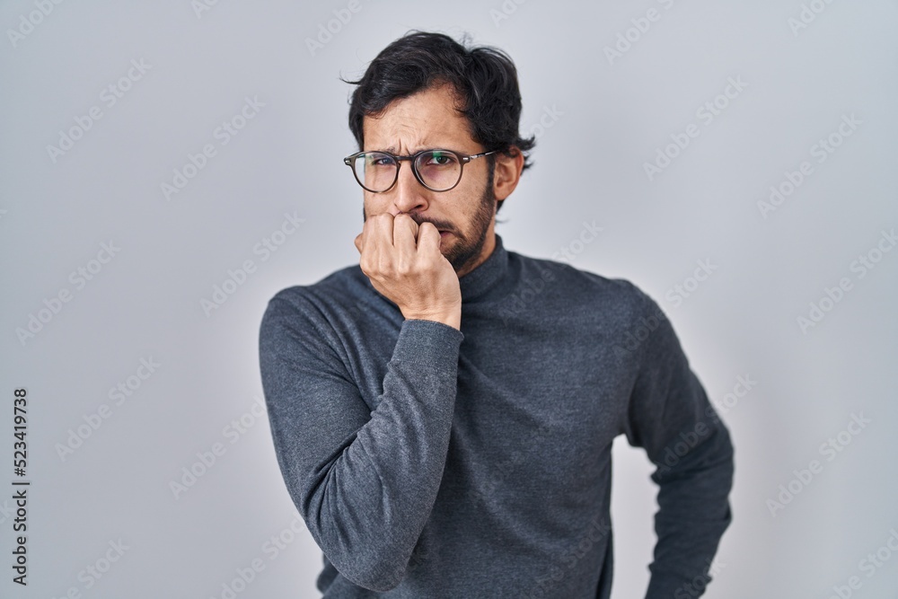 Handsome latin man standing over isolated background looking stressed and nervous with hands on mouth biting nails. anxiety problem. Handsome latin man standing over isolated background looking stressed and nervous with hands on mouth biting nails. anxiety problem.