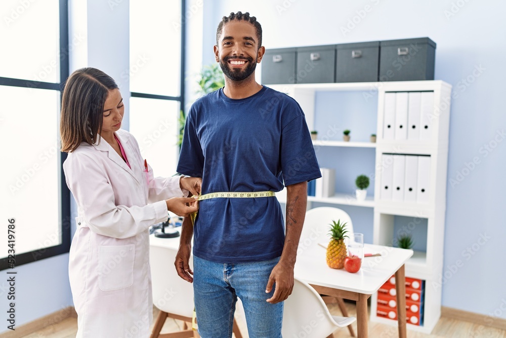 Obraz premium Dietitian at the clinic using tape measure measuring waist of client looking positive and happy standing and smiling with a confident smile showing teeth