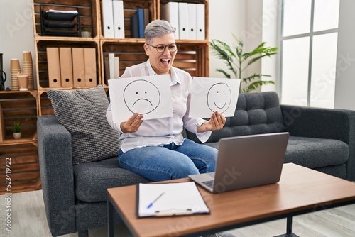 Middle age woman working on depression holding sad to happy emotion paper smiling and laughing hard out loud because funny crazy joke.